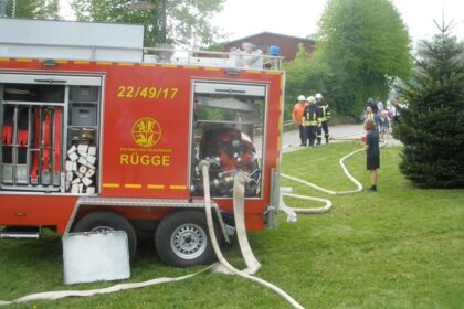Geöffneter roter Feuerwehranhänger mit Pumpe, Schläuche liegen auf der Wiese, im Hintergrund Feuerwehrleute und Kinder