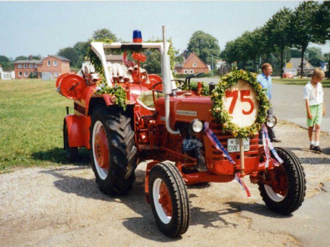 Geschmückter roter Traktor mit Tragkraftspritze und einem Schild mit der Zahl 75 vor einer Dorfstraße