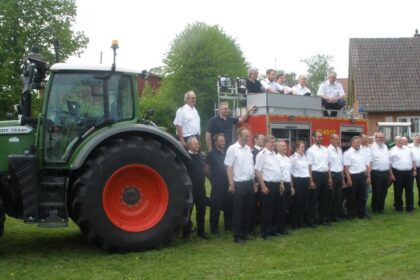 Feuerwehrleute in weißen Uniformhemden stehen in einer Reihe vor einem roten Feuerwehranhänger und einem Traktor